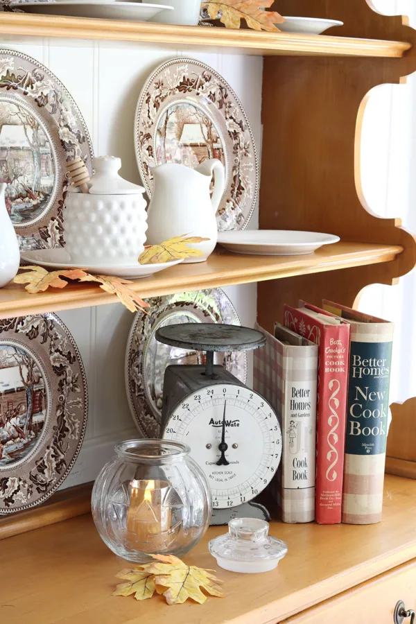 Neutral and orange fall decorations in the kitchen featuring a hutch filled with vintage autumn colored dishes and autumn leaves.