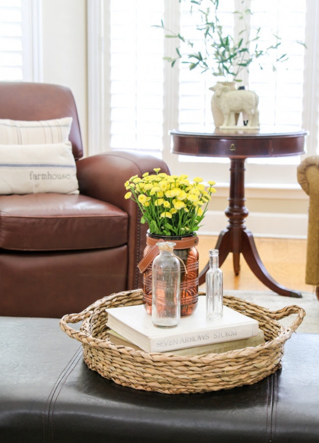 Fall basket tray idea. Sitting on an ottoman is a round basket tray with a stack of books. Sitting on the books is a amber glass jar with fresh fall flowers in it. Then two vintage glass clear jars beside it.