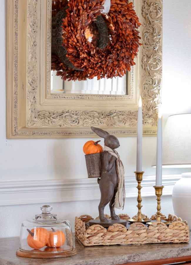 Fall entryway tray idea is a scallop basket tray with two brass candlestick holder with taper candles. A figurine of a bunny standing with a basket backpack with a bright orange mini pumpkin in is basket. Next to the basket is a cloche with two bright orange pumpkins in it.