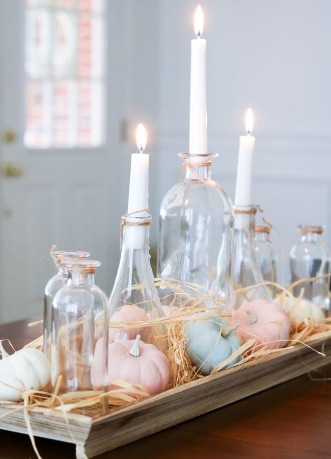 Easy fall tray centerpiece idea. On a dining table is a wooden long tray with clear jars of different heights with taper candles in them then on the tray is raffia and mini pumpkins painted in pastel colors for fall.