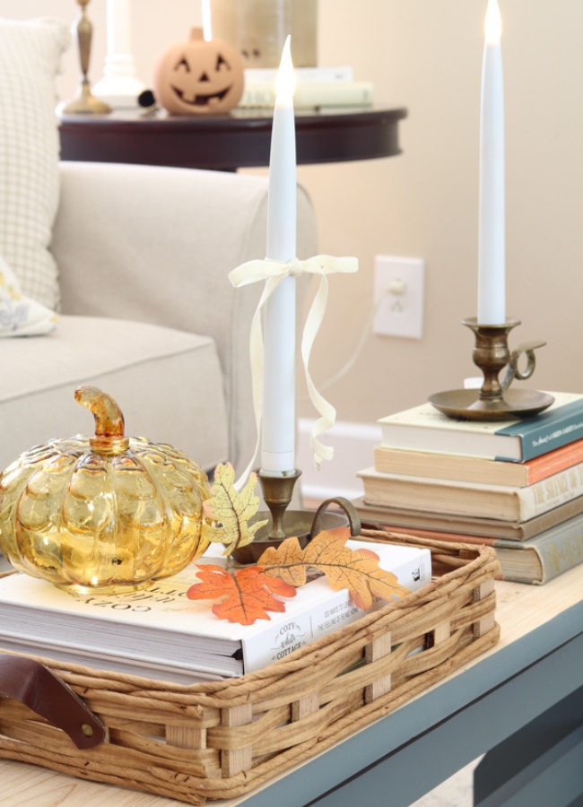 Fall coffee table tray idea. A basket tray with books on the bottom, brass candle stick holder with taper candle, glass pumpkin with fairy lights inside and a sprig of leaves. On the right is a stack of books with another candle on it and this all is sitting on a bench coffee table.