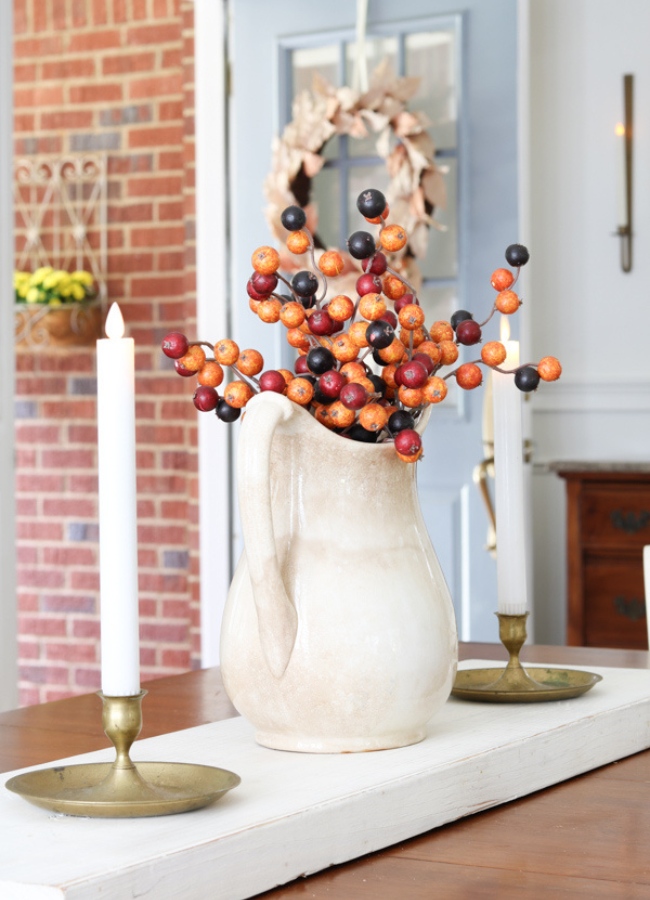 This fall tray decor idea example is to show how a piece of wood can act as a fall tray. It is a simple piece of wood painted with white chalk paint. On the wood board tray is a vintage ironstone pitcher with autumn berries in it and on each side of the pitcher is a brass candlestick holder with taper candles.