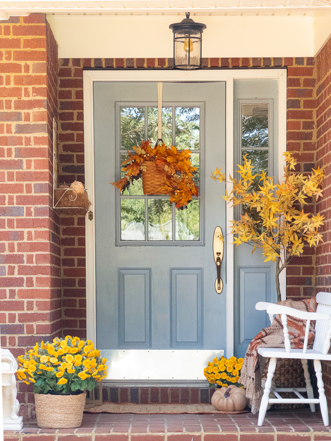 Fall front porch ideas.  Brick house with blue door.  Basket hanging on door filled with autumn leaves of orange.  Tall maple tree in right corner with yellow leaves.  White bench on the right with a autumn colored blankets draped on it.  Faux yellow mums on the floor with faux pumpkins.  On the left two Dollar Tree lanterns, another faux mum and a white dog statue.