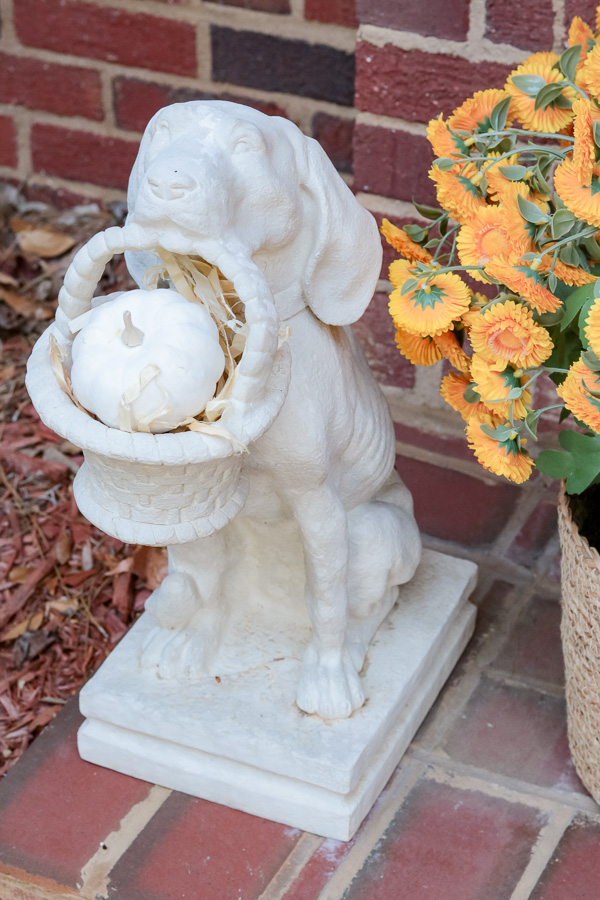 Dog statue holding a basket and inside the basket is hay and a white painted pumpkin.