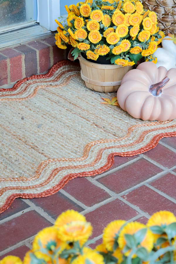 Jute scallop rug on a brick floor.  The rug is tirmmed in different shades of orange.  In the corner are faux pumpkins and a faux mum in a bushel basket.