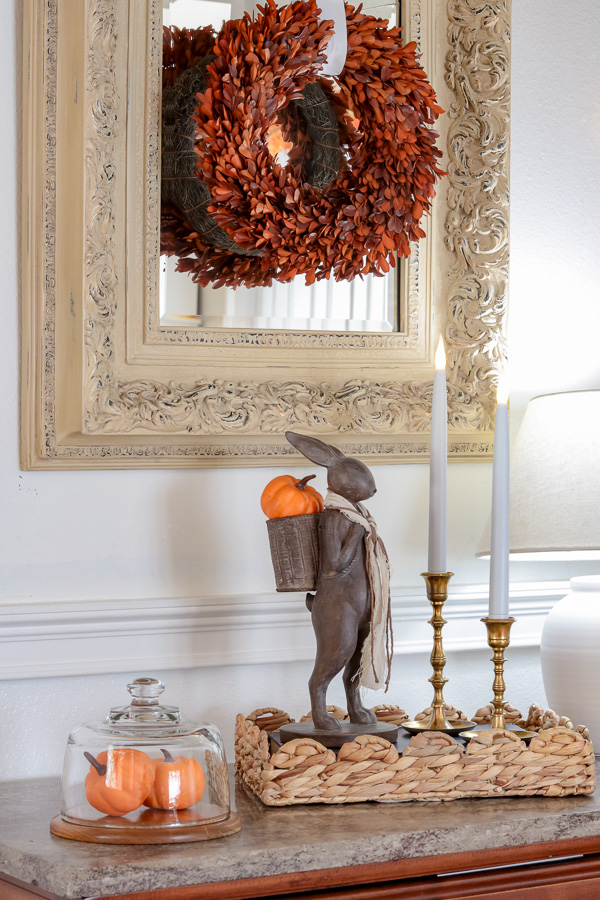 Entryway fall decorations.  Mini pumpkins under cloche, Standing rabbit with mini pumpkin in his backpack, candles, on a book in a basket tray.  Haning on a mirror is a burnt orange boxwood wreath.