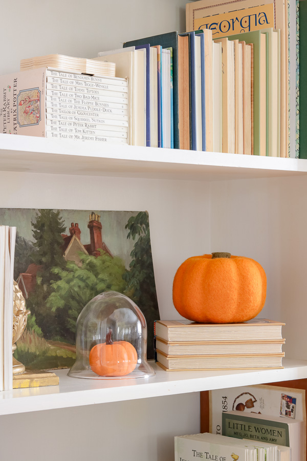 Fall decorations on a bookcase with faux pumpkins.  One sitting on a stack of book while another tiny pumpkin is under a cloche.
