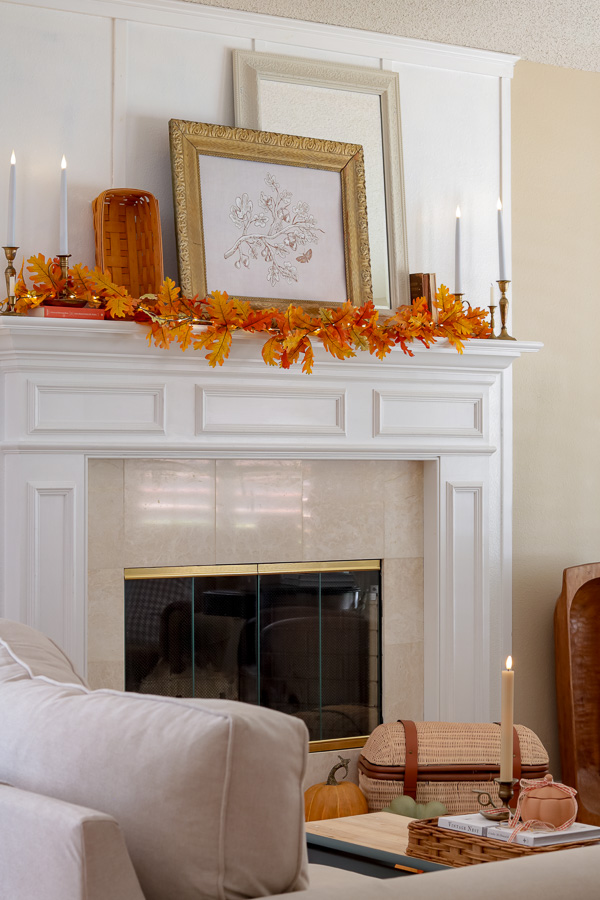 Fireplace mantel decorated for fall.  Layered mirror and fall botanical art, autumn leaf garland across mantel, brass candlesticks with battery powered candles, Longaberger basket, old books.