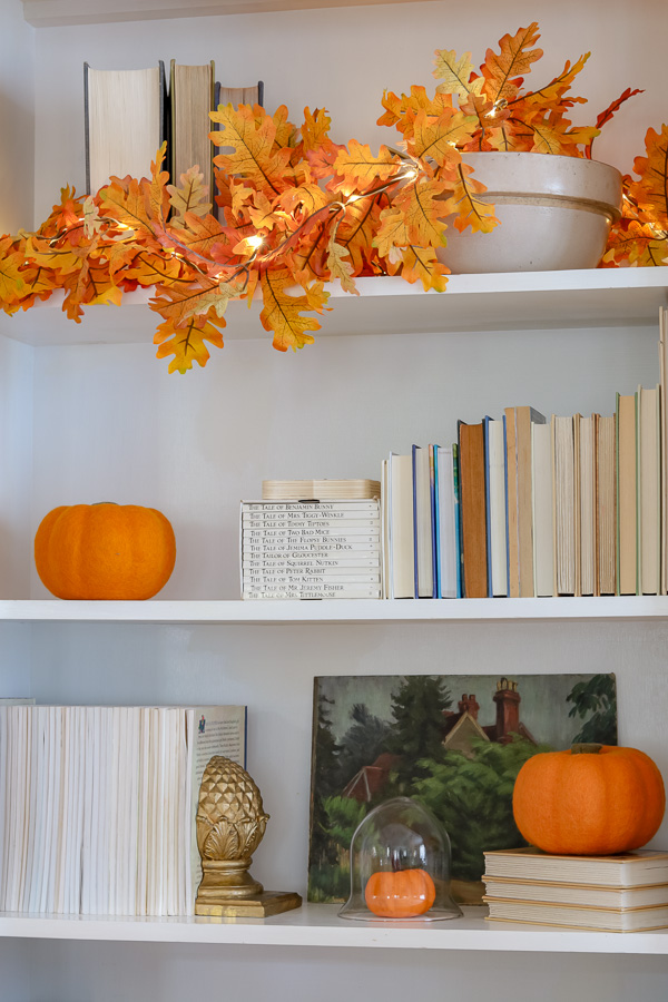 Fall decorations on a bookcase with faux pumpkins and autumn leaf garland.