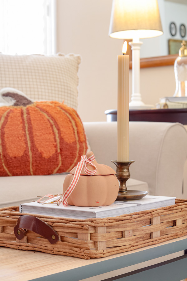 Basket tray with books stacked in it and a terracotta pumpkin jar on it with gingham bow tied to the stem.  Vintage brass candlestick holder with tan battery powered taper candle.  Pumpkin pillow in the back ground on sofa.