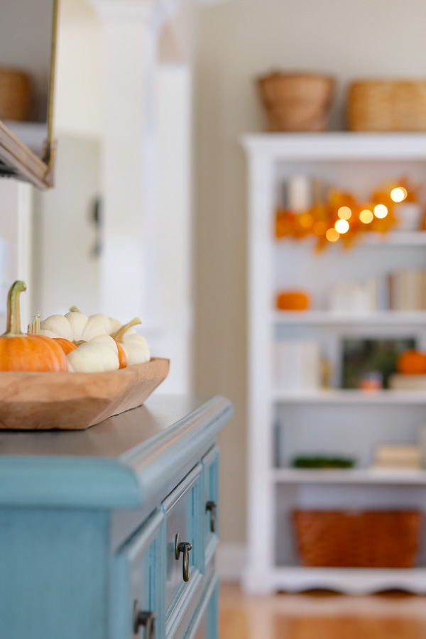 Dough bowl filled with white and orange mini pumpkins on a blue cabinet and a bookcase in the back ground decorated with fall decor.