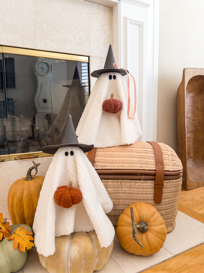 Cute sherpa ghost on a wicker basket runk with faux pumpkins and autumn leaves on a fireplace hearth.
