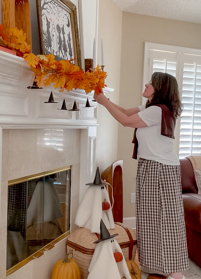 Hanging a mini paper witch hat garland on a mantel layered with a fall leaf garland for cozy Halloween decor.