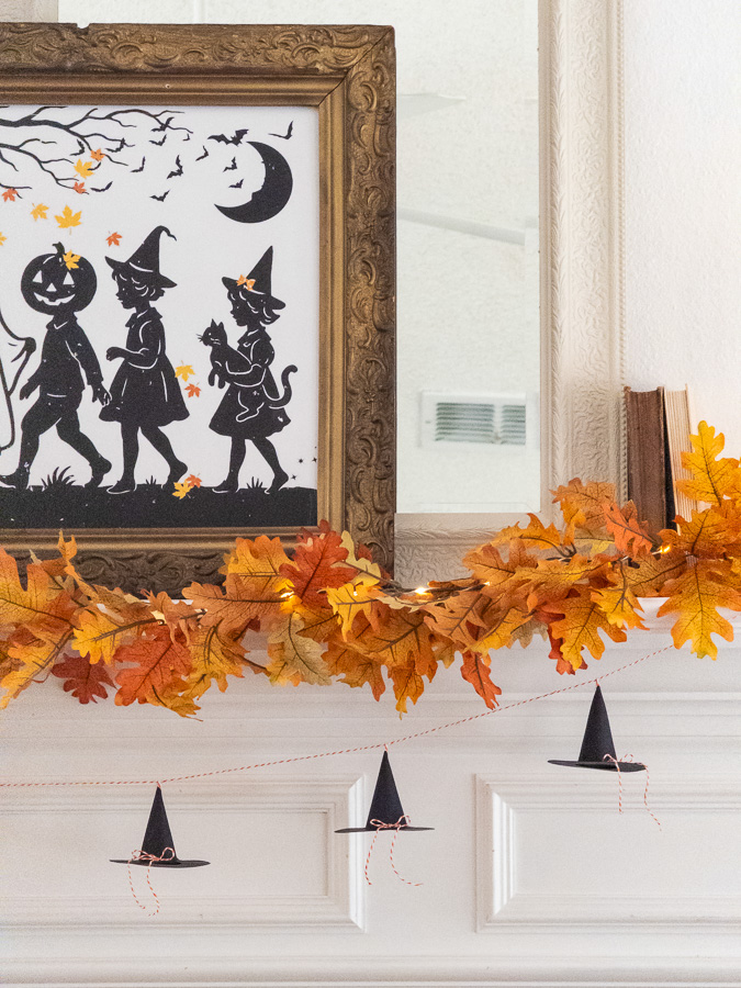 Shelf with autumn leaf garland, mini paper witch hat garland and art silhouette of kids trick or treating in costumes