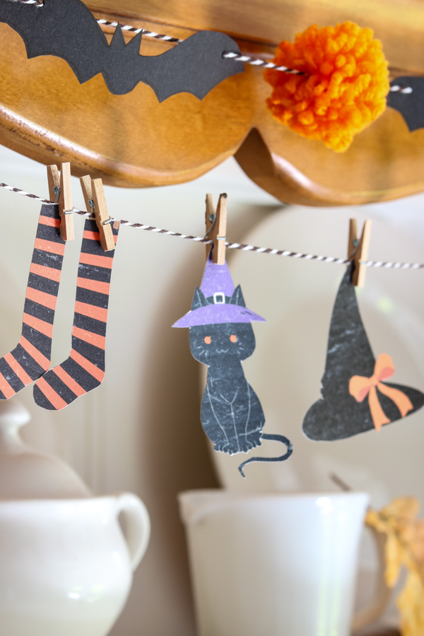 Close-up of a witch’s clothesline garland with tiny paper witch clothes and black cat hanging above a black bat and orange pom pom garland, layered Halloween garland DIY.