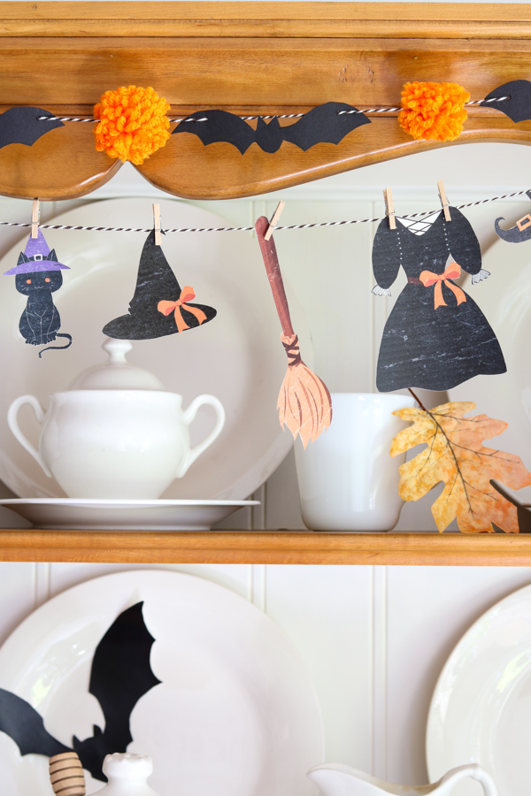 Close-up of a witch’s clothesline garland with tiny paper witch clothes and black cat hanging above a black bat and orange pom pom garland, layered Halloween garland DIY.
