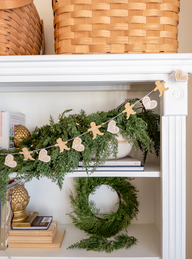Handmade cookie cutter gingerbread man, woman and heart garland in front of a cypress garland filled with lights on a bookcase