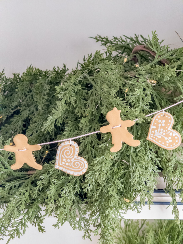 Handmade cookie cutter gingerbread man, woman and heart garland in front of a cypress garland filled with lights on a bookcase