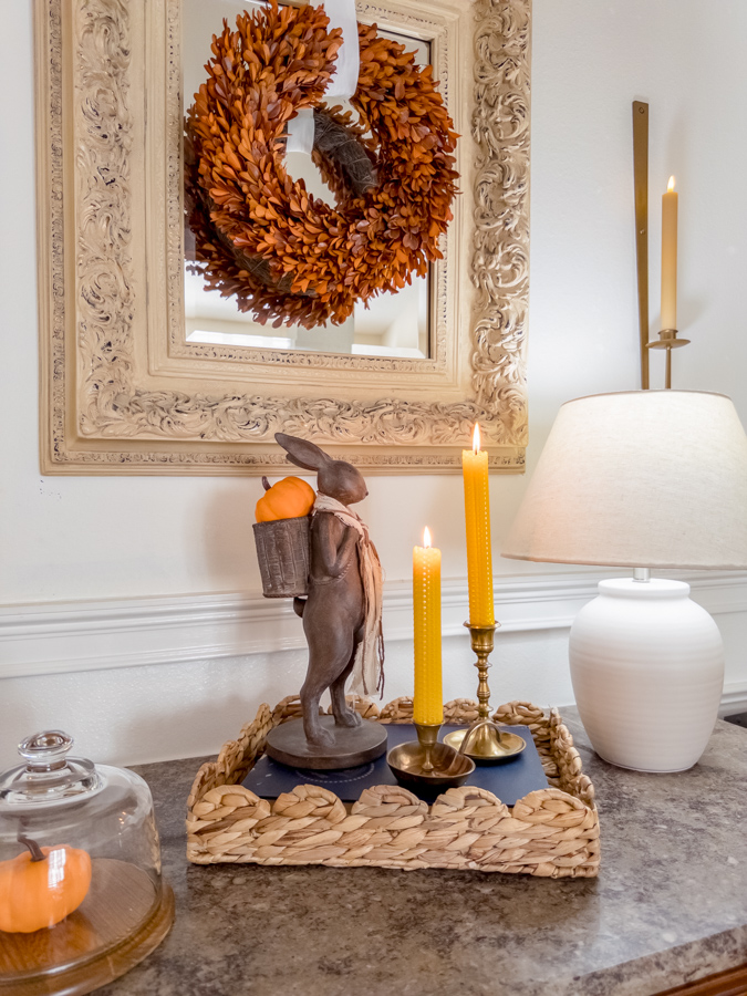 Entryway decorating for fall with a lamp, basket tray with book inside and two brass candle stick holder with rolled beeswax candles, a bunny with basket and a pumkin in his basket.