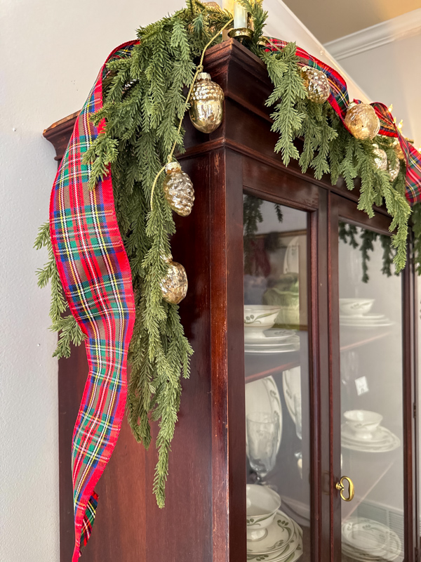 Layered greenery, tartan ribbon, pinecone garland and Christmsa lights on a dark wood china cabinet