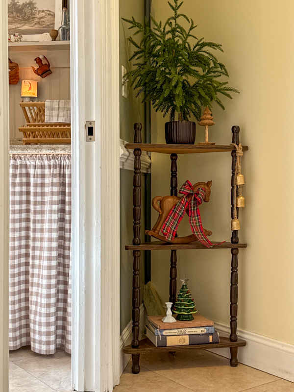 Corner next to kichen with vintage shelves and Christmas decorations.  Small tree, bells hanging on furniture, wood rocking horse with plaid ribbon bow, ceramic Christmas tree sitting on a stack of books.