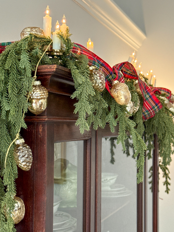 Greenery garland, plaid ribbon, pinecone garland and Christmas lights on a china cabinet