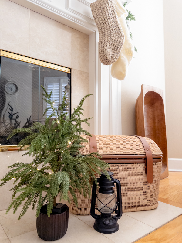 Fireplace hearth decorated for Christmas with a basket trunk, large wood dough bowl, old barn lantern and small Christmas tree.