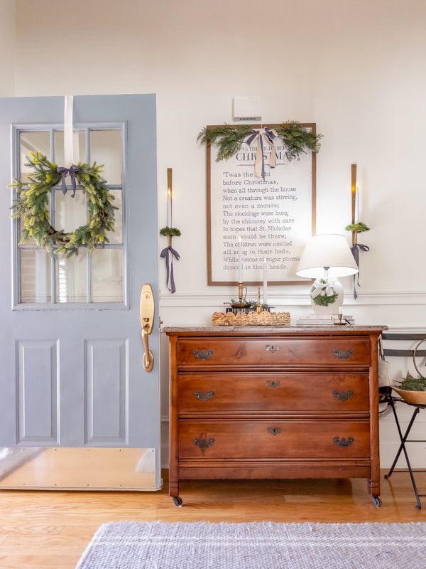 Entryway decorated for Christmas inspired by Little House on the Prairie. Blue front door with greenery wreath with blue bow. Twas the Night Before Christmas bookpage art over chest of drawer. Greenery on top of art with bow. Wall scones on each side tied with blue bows and greenery. Basket tray on cabinet with brass candles, books and greenery. White lamp with wreath and ticking bow.