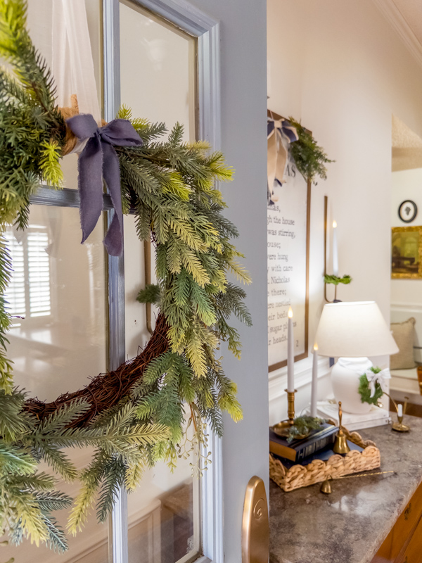 Entryway decorated for Christmas inspired by Little House on the Prairie. Blue front door with greenery wreath with blue bow. Twas the Night Before Christmas bookpage art over chest of drawer. Greenery on top of art with bow. Wall scones on each side tied with blue bows and greenery. Basket tray on cabinet with brass candles, books and greenery. White lamp with wreath and ticking bow.