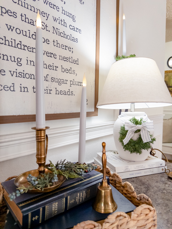 Entryway decorated for Christmas inspired by Little House on the Prairie. Basket tray on cabinet with brass candles, books and greenery. White lamp with wreath and ticking bow. Modern prarie look.