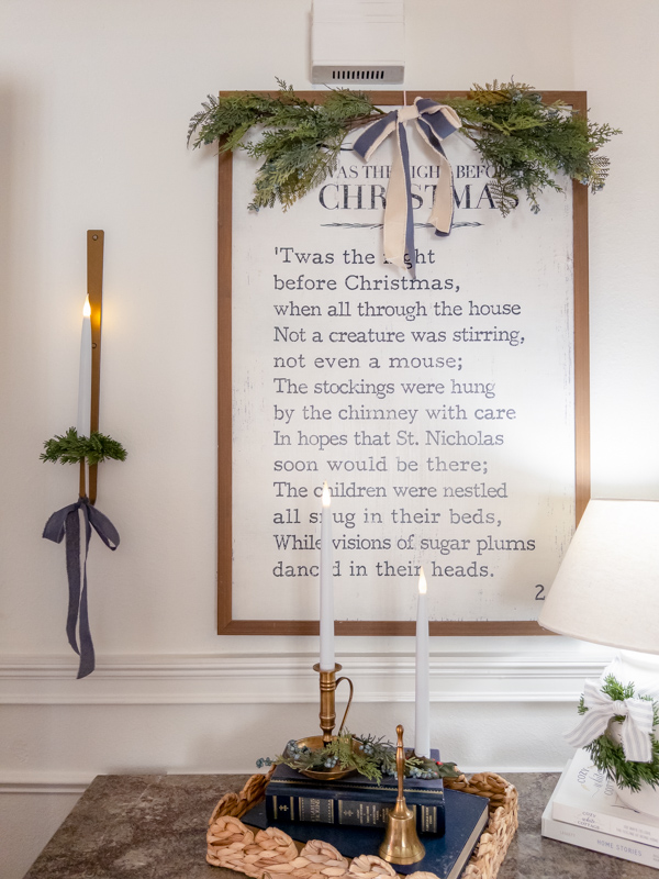 Entryway decorated for Christmas inspired by Little House on the Prairie. Twas the Night Before Christmas bookpage art over chest of drawer. Greenery on top of art with bow. Wall scones on each side tied with blue bows and greenery. Basket tray on cabinet with brass candles and bell, books and greenery. White lamp with wreath and ticking bow.