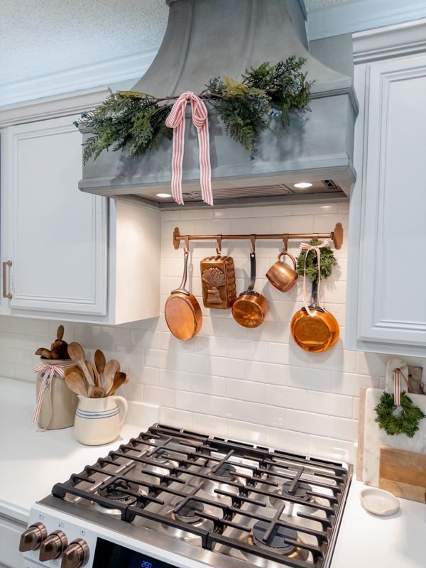 Blue range hood decorated for Christmas with greenery tied with red and white ribbon. Copper pans hanging over the stove with mini wreath with red and white ribbon. Crocks on one side of hte stove filled with old wooden spoons and cutting boards on other side with mini wreath.