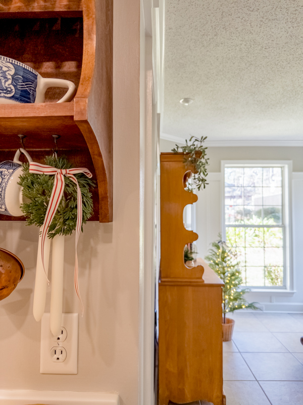 View from kitchen to sitting room. Small wall hutch with mini wreath with red and white bow, Currier and Ives tea cup and hanging candles.