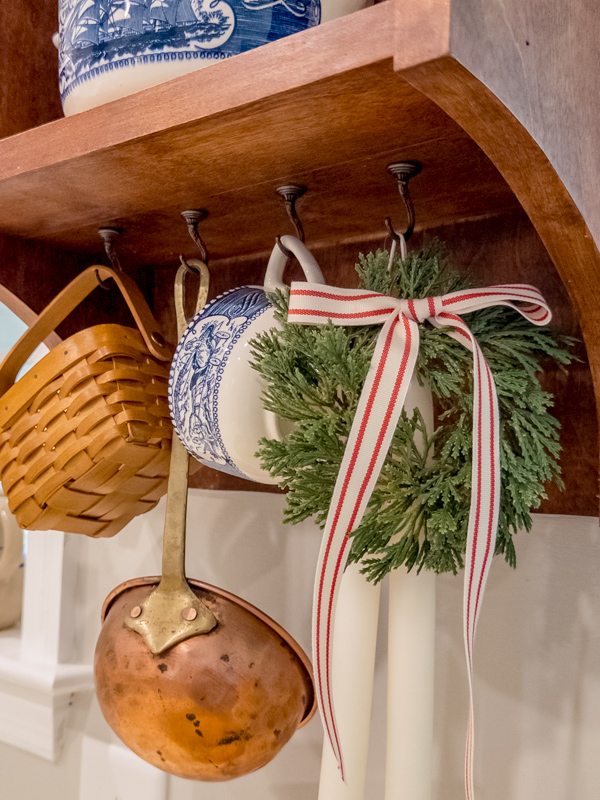 Small wall hutch with hooks decorated for Christmas. Hanging on the hooks is a basket, old copper ladel like they would have used on the prairie, blue and white teacup, hanging candles and mini wreath with red and white ribbon.