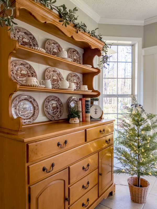 Hutch decorated for Christmas with brown and blue Currier and Ives style dishes and vintage Santa mugs, old scale, garland across top, vintage cookbooks and small Christmas tree topped with ribbon bow.