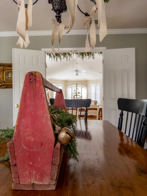 Formal dining room Little House on the Prairie inspired table centerpiece. Old wood toolbox like Charles Ingalls, filled with greenery and rustic jingle bells, Chandelier with off white cotton ribbon bows.