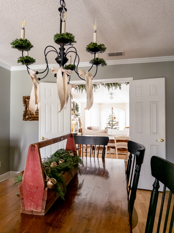 Dining room inspired by Little House with chandelier with greenery and cotton bows and table centerpiece of an old wooden toolbox filled with greenery and bows.