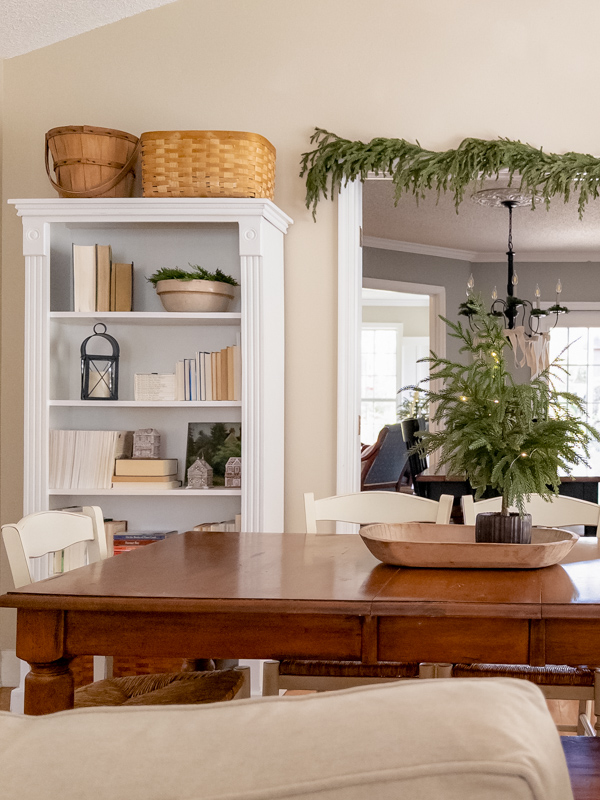 View from informl dining room into living room with a garland over the door and bookcase on one side decorated with vintage decor - lanterns, crock bowls with greenery, gingerbread houses. On the table is a wood dough bowl tray with a small Christmas tree.