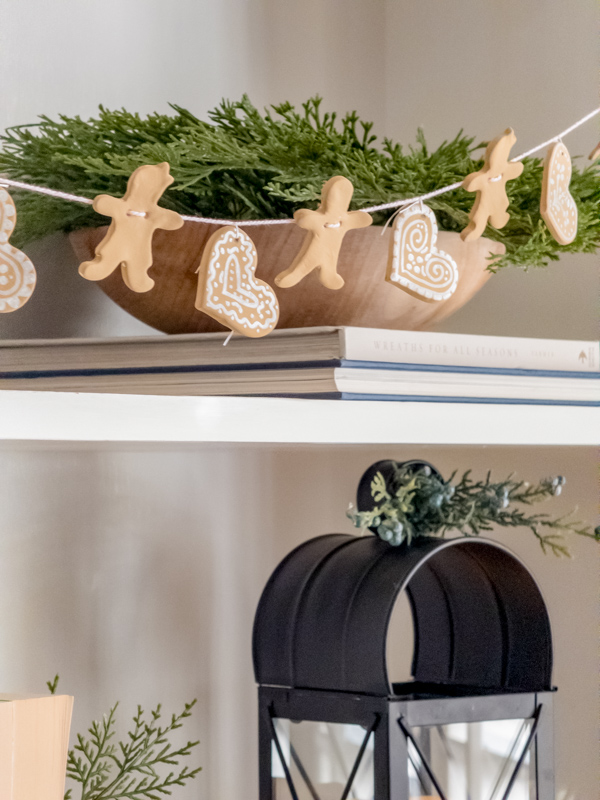 Gingerbread men and heart garland made from air dry clay for Christmas hanging on a bookcase in front of a wood bowl with garland and books.