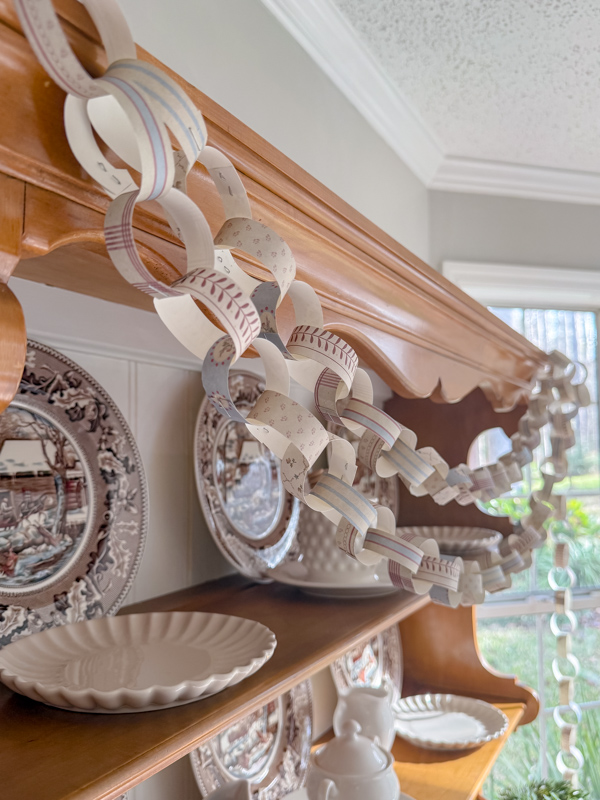 Hutch decorated for winter with old winter scene dishes Currier & Ives in browns and blues. And a cozy cottage pattern paper chain garland draped across the hutch.