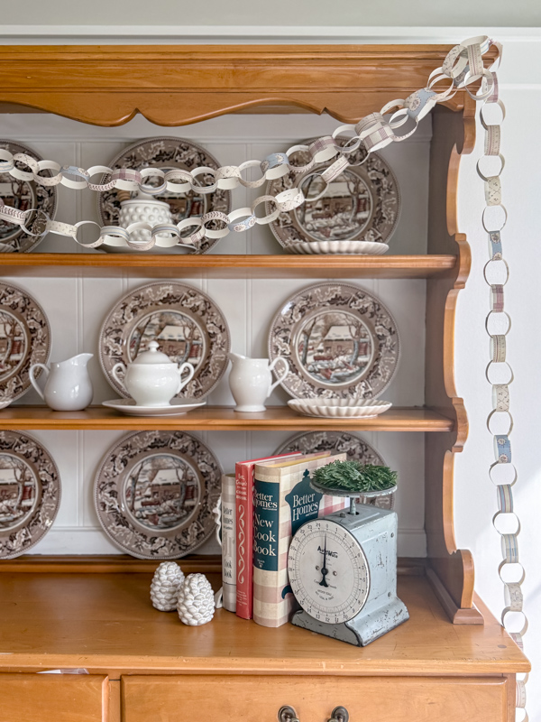 Hutch decorated for winter with old winter scene dishes Currier & Ives in browns and blues. And a cozy cottage pattern paper chain garland draped across the hutch.
