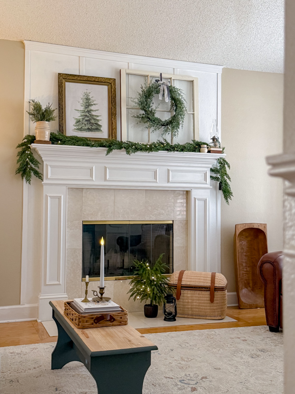 Winter decorating ideas living room fireplace mantel dressed with cedar garland, fir tree art, old window with cedar wreath, vintage crock with sprigs of greenery, chamber lamp sitting on stack of old books.  On the heart is a basket chest for wood, mini tree with fairy lights and old barn lantern.