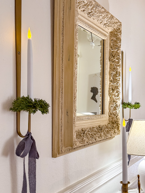 Vintage ornate mirror and candle sconces on a wall dressed with tiny cedar wreaths on the candle holder and blue ribbon tied in a droppy bow.