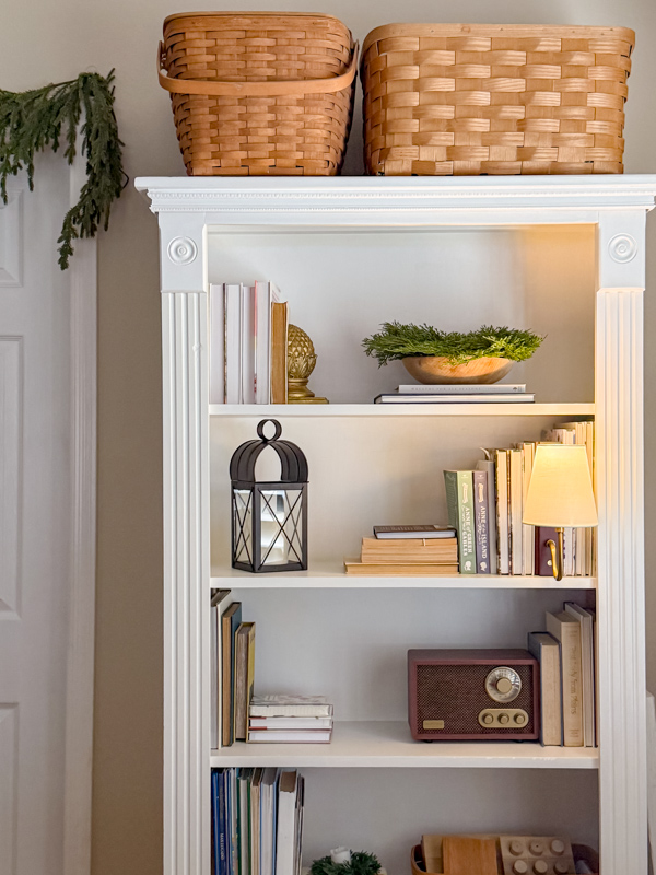 Bookcase decorated with non Christmas winter decor. Cedar wreaths on bowls, book lamp and old vintage radio which is actually a new Joanna Gaines radio made to look old.