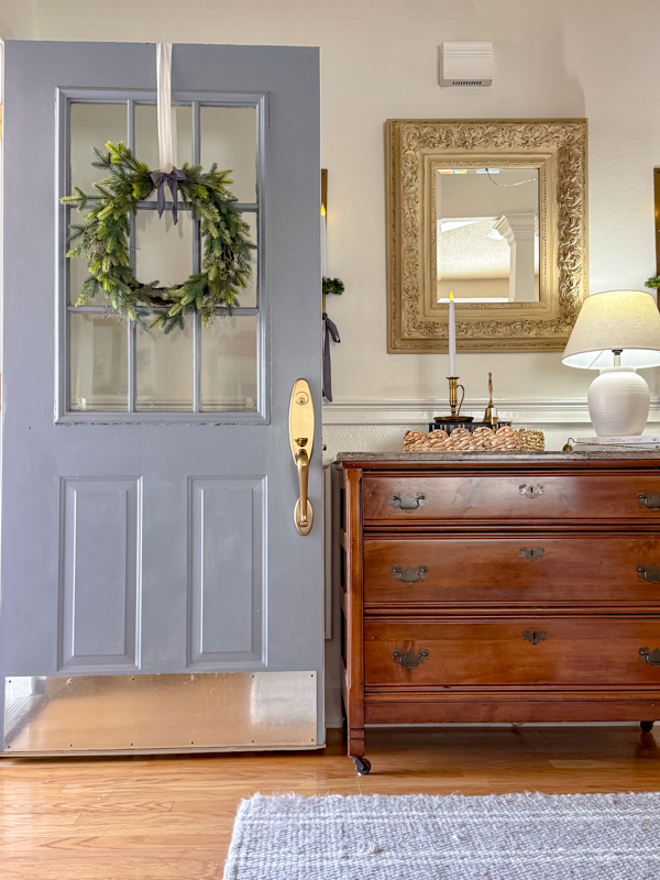 Entryway winter decor ideas. Blue door with greenery wreath tied with blue ribbon. Chest decorated with basket tray, books and candles, and wall candle sconces has a mini wreath with blue ribbon tied in a droopy bow.