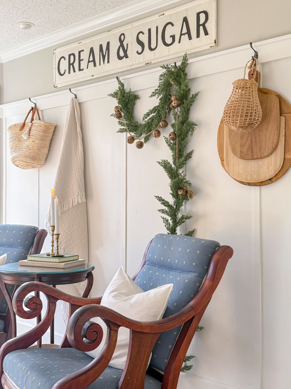 Wall full of hooks decorated for winter with a garland with rustic bells and fairy lights. Basket, blanket and wood cutting and break boards layered with a garlic basket