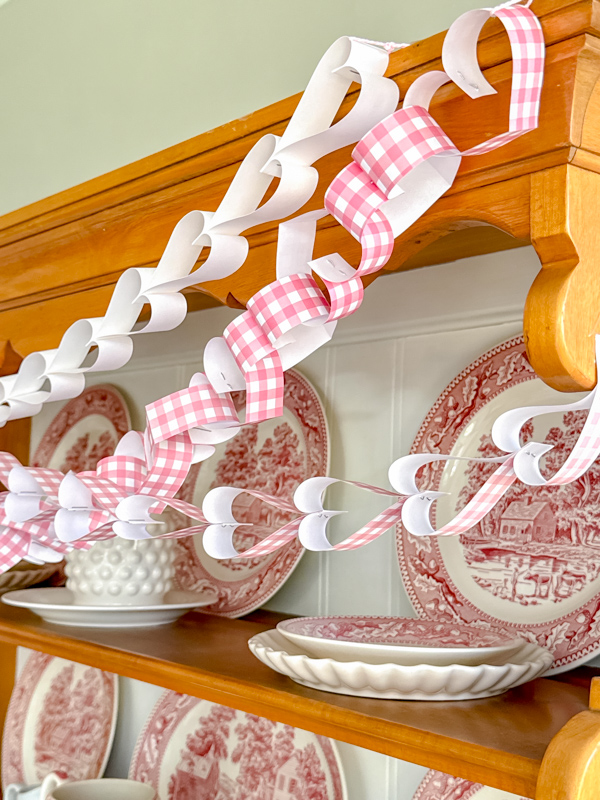 DIY paper heart chain garland.  Three garlands drapped on a hutch.  One is a all white while the other two are pink and white gingham.