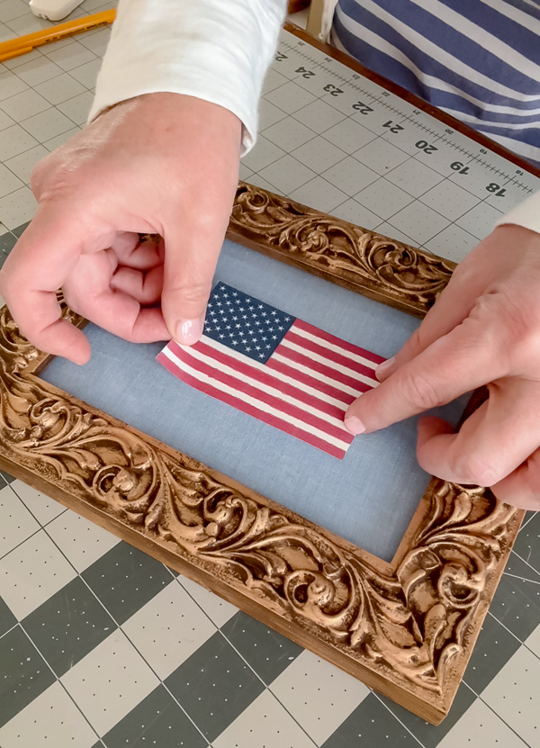 Placing a printed American flag onto a chambray fabric background inside a vintage gold ornate frame for DIY framed American flag art