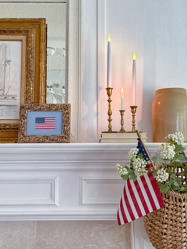 Patriotic fireplace mantel decorated with DIY framed American flag art on chambray fabric, brass candlesticks, a sailboat printable, and a wicker basket with white flowers and a stick flag for 4th of July home decor