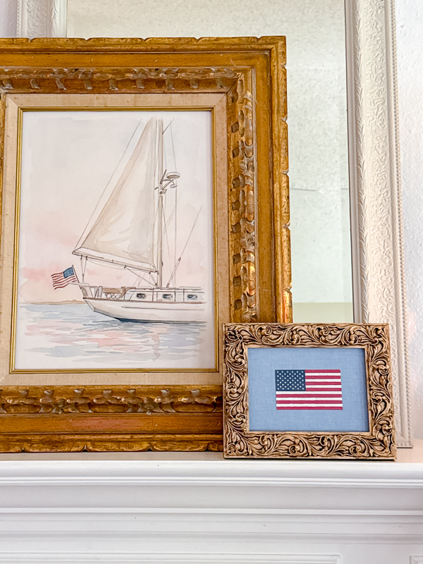 Fireplace mantel styled with a large sixteen by twenty watercolor sailboat print from Walgreens and a smaller American flag printed on watercolor canvas paper at home both framed in vintage thrifted gold ornate frames with the flag displayed on a chambray fabric background